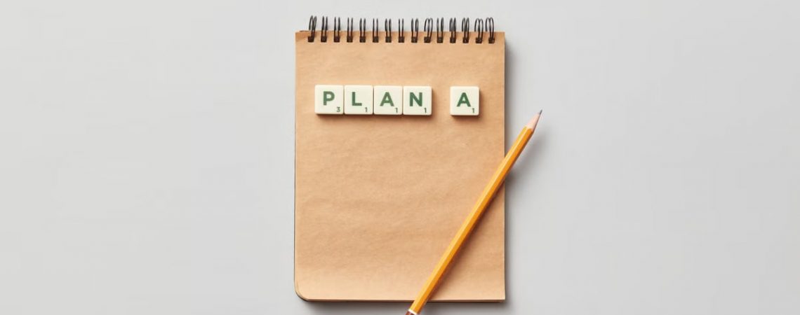 Top view of a notepad with a pencil and scrabble tiles spelling 'PLAN A' on a gray background.